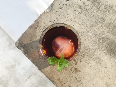 A glass of dark brown iced beverage with a large ice cube and a sprig of fresh mint is placed on a textured stone surface. The lighting creates a contrast between the cool drink and the rough, light-colored texture of the surface.