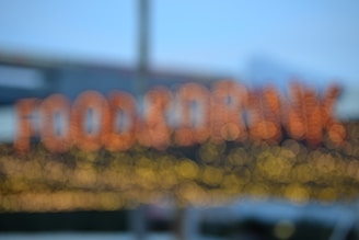 A blurred image with out-of-focus lights forming the words 'FOOD & DRINK' in a bokeh style. The background is soft, with hints of a blue sky and some structural elements possibly from a building.