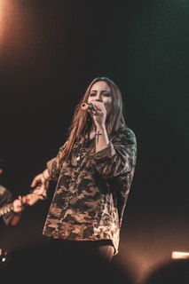 A person with long hair is singing into a microphone while wearing a camouflage patterned shirt. The background is dark, with a subtle light creating a warm ambiance. Focus is on the singer, with a blurred instrument visible in the background.