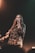 A person with long hair is singing into a microphone while wearing a camouflage patterned shirt. The background is dark, with a subtle light creating a warm ambiance. Focus is on the singer, with a blurred instrument visible in the background.