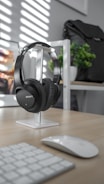A pair of black, over-ear Sony headphones is neatly placed on a white stand on a wooden desk. In the background, a black backpack is partially visible on a shelf, next to a small green plant. A wireless mouse and keyboard are on the desk, suggesting a clean and organized workspace. Sunlight streams through blinds, casting striped shadows across the scene.