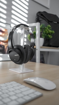 A pair of black, over-ear Sony headphones is neatly placed on a white stand on a wooden desk. In the background, a black backpack is partially visible on a shelf, next to a small green plant. A wireless mouse and keyboard are on the desk, suggesting a clean and organized workspace. Sunlight streams through blinds, casting striped shadows across the scene.