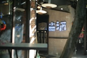 A dimly lit entrance to a restaurant or bar with a rustic interior featuring brick walls and soft lighting. A neon sign with Asian characters and the text 'Mochu Kitori' is mounted on the wall. There are reflections on glass surfaces creating a layered, slightly hazy effect.