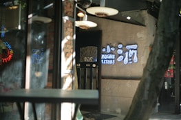 A dimly lit entrance to a restaurant or bar with a rustic interior featuring brick walls and soft lighting. A neon sign with Asian characters and the text 'Mochu Kitori' is mounted on the wall. There are reflections on glass surfaces creating a layered, slightly hazy effect.