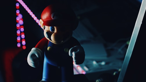 A figurine resembling a video game character stands in the foreground, partially illuminated by colorful, circular lights in the background. The setting is dark with muted colors, creating a sense of mystery. The character wears a red cap and blue overalls, holding a determined expression.