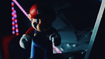 A figurine resembling a video game character stands in the foreground, partially illuminated by colorful, circular lights in the background. The setting is dark with muted colors, creating a sense of mystery. The character wears a red cap and blue overalls, holding a determined expression.