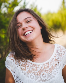A young woman with brown hair is laughing joyfully, wearing a white lace top. Her eyes are closed and her hair is slightly tousled by the wind, adding to a carefree and happy atmosphere. The background is softly blurred with greenery, suggesting an outdoor setting.