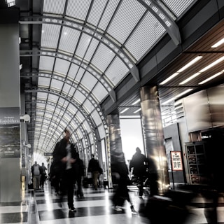 A busy and modern indoor public space with a high arched ceiling made of glass and metal. People are walking, some in motion blur, indicating movement and hustle. There are advertisements on the walls, including one for Hilton and a sign offering a 30% discount. The lighting is bright, and the space feels expansive and contemporary.