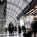 A busy and modern indoor public space with a high arched ceiling made of glass and metal. People are walking, some in motion blur, indicating movement and hustle. There are advertisements on the walls, including one for Hilton and a sign offering a 30% discount. The lighting is bright, and the space feels expansive and contemporary.