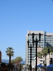 A cityscape featuring a tall modern building with the Adobe logo on top. The foreground has several black streetlights and palm trees lining the street. The clear blue sky serves as a backdrop.