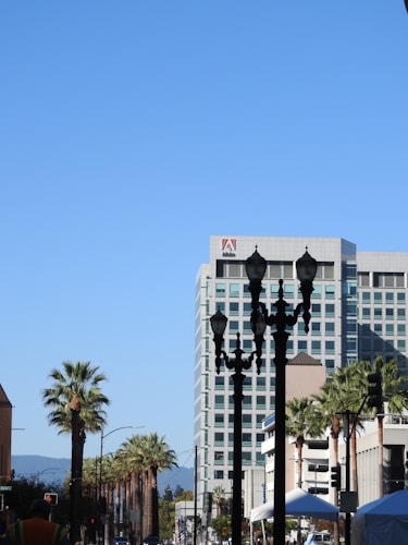 A cityscape featuring a tall modern building with the Adobe logo on top. The foreground has several black streetlights and palm trees lining the street. The clear blue sky serves as a backdrop.