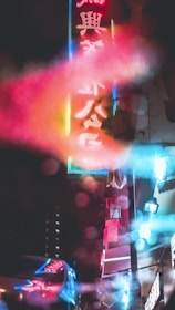 A blurred view of neon signs in an urban street at night. The signs display Chinese characters, and the image has a dreamy, surreal feel due to the colorful light reflections. Buildings and additional signage are partially visible in the background.