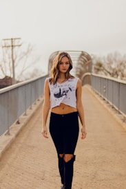 A young woman with long hair walks confidently down a dirt path on a pedestrian bridge. She is wearing a white crop top with 'Los Angeles' printed on it and black ripped jeans. The bridge is enclosed with metal railings, and the background is slightly blurred with leafless trees.