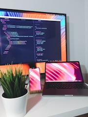 A modern workstation setup with a large computer monitor displaying code, a laptop, and a smartphone with matching colorful backgrounds. A potted green plant is placed on the desk, adding a touch of nature to the otherwise tech-focused environment.