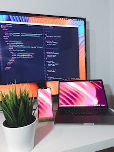 A modern workstation setup with a large computer monitor displaying code, a laptop, and a smartphone with matching colorful backgrounds. A potted green plant is placed on the desk, adding a touch of nature to the otherwise tech-focused environment.