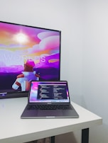 A modern workspace featuring a MacBook Pro placed on a white desk. The laptop screen displays a programming code editor with multiple lines of code. In the background, a television screen shows an animated scene with vibrant colors, including a character in futuristic armor, against a colorful sky with text partially visible.