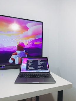 A modern workspace featuring a MacBook Pro placed on a white desk. The laptop screen displays a programming code editor with multiple lines of code. In the background, a television screen shows an animated scene with vibrant colors, including a character in futuristic armor, against a colorful sky with text partially visible.