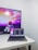 A modern workspace featuring a MacBook Pro placed on a white desk. The laptop screen displays a programming code editor with multiple lines of code. In the background, a television screen shows an animated scene with vibrant colors, including a character in futuristic armor, against a colorful sky with text partially visible.