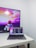 A modern workspace featuring a MacBook Pro placed on a white desk. The laptop screen displays a programming code editor with multiple lines of code. In the background, a television screen shows an animated scene with vibrant colors, including a character in futuristic armor, against a colorful sky with text partially visible.