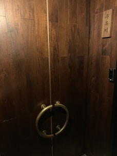 A set of large wooden double doors with a dark brown finish, featuring circular metal handles. There is a wooden plaque with Korean characters on the right side.
