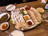A platter of food including sliced boiled pork, sliced octopus, and various side dishes such as kimchi and garlic. There are dipping sauces in small bowls. The table is set with chopsticks, a bottle, and side plates containing different condiments and dishes.