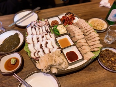 A platter of food including sliced boiled pork, sliced octopus, and various side dishes such as kimchi and garlic. There are dipping sauces in small bowls. The table is set with chopsticks, a bottle, and side plates containing different condiments and dishes.