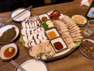 A platter of food including sliced boiled pork, sliced octopus, and various side dishes such as kimchi and garlic. There are dipping sauces in small bowls. The table is set with chopsticks, a bottle, and side plates containing different condiments and dishes.