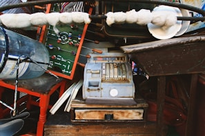 An eclectic collection of vintage and miscellaneous items is densely arranged. A prominent piece is an old cash register with visible numbered keys and a weathered appearance. A foosball table can be seen, notably its miniature players and green playing field. Surrounding these are a red-painted chair, a large glass bottle, a bundle of cotton or wool wrapped on rods, and various wooden and metal objects.