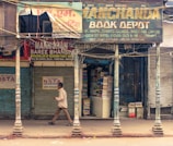 A street scene features a pedestrian walking past a row of closed shops. One shop, Manchanda Book Depot, is open, filled with piles of books stacked inside. The exterior of the building is weathered, with peeling paint and old signage in multiple languages. Metal shutters are visible, some painted with advertisements, and the overall atmosphere suggests a bustling marketplace.