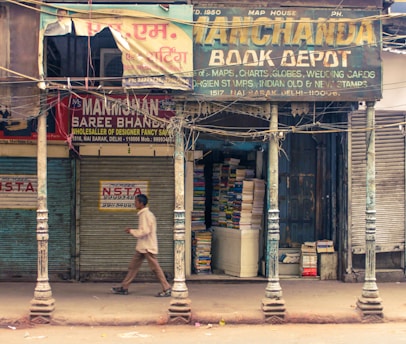 A street scene features a pedestrian walking past a row of closed shops. One shop, Manchanda Book Depot, is open, filled with piles of books stacked inside. The exterior of the building is weathered, with peeling paint and old signage in multiple languages. Metal shutters are visible, some painted with advertisements, and the overall atmosphere suggests a bustling marketplace.