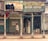 A street scene features a pedestrian walking past a row of closed shops. One shop, Manchanda Book Depot, is open, filled with piles of books stacked inside. The exterior of the building is weathered, with peeling paint and old signage in multiple languages. Metal shutters are visible, some painted with advertisements, and the overall atmosphere suggests a bustling marketplace.