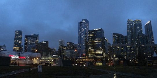 A nighttime cityscape featuring tall, illuminated skyscrapers against a dark, cloudy sky. The skyline includes a variety of building styles, with both rectangular and curved towers. Ground level structures are visible, with one prominently displaying the word 'Canada'. Sparse lighting and shadows create a dramatic effect.