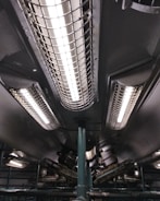 A series of industrial fluorescent lights are encased in protective grids, mounted on a dark ceiling with visible pipes and mechanical components.