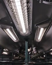 A series of industrial fluorescent lights are encased in protective grids, mounted on a dark ceiling with visible pipes and mechanical components.