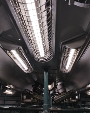 A series of industrial fluorescent lights are encased in protective grids, mounted on a dark ceiling with visible pipes and mechanical components.