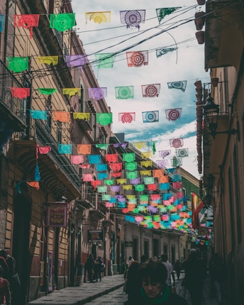 An urban street is adorned with colorful papel picado banners hanging above, providing a festive atmosphere amidst a historical architectural setting. The buildings are old with balconies and various signs in Spanish. People are walking along the street, suggesting a lively, busy street scene.