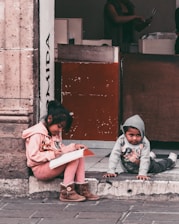 Two children are sitting on the sidewalk, with one child writing in a notebook and the other crawling on the ground. They are positioned in front of a building entrance that features a textured stone wall and a wooden door. The scene appears casual and everyday, capturing a moment of concentration and play.