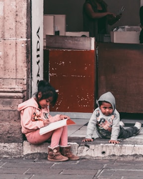 Two children are sitting on the sidewalk, with one child writing in a notebook and the other crawling on the ground. They are positioned in front of a building entrance that features a textured stone wall and a wooden door. The scene appears casual and everyday, capturing a moment of concentration and play.