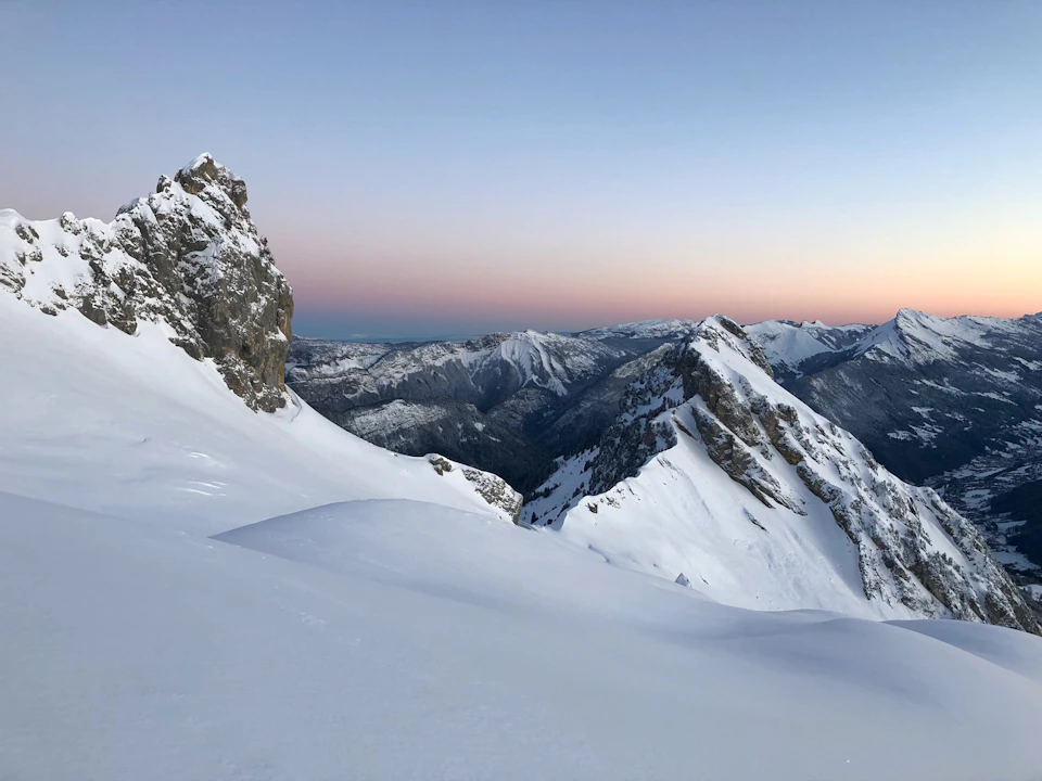 The image depicts a breathtaking view of a snow-covered mountain range at sunset. The mountains, blanketed in pristine white snow, rise majestically against the sky, their peaks dusted with hues of orange and pink. The sun is setting behind these majestic peaks, casting a warm glow over the scene. The perspective of the image is from a high vantage point, looking down upon the mountains, which enhances the grandeur of the landscape.