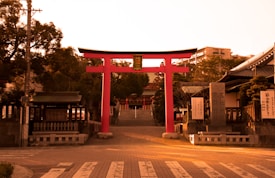 A large red torii gate stands prominently in front of a pathway leading to a traditional Japanese shrine. The gate is flanked by lanterns and buildings with tiled roofs on either side. Trees and lush foliage partially conceal the structures behind the gate. The pathway is lined with stone lanterns and the setting sun casts a warm, golden hue over the whole scene.