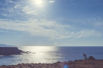 A scenic landscape featuring a rugged coastline under a clear sky. The sunlight reflects off the ocean, creating a shimmering effect on the water. A person is seen on the right setting up a camera on a tripod, capturing the beauty of the scene.
