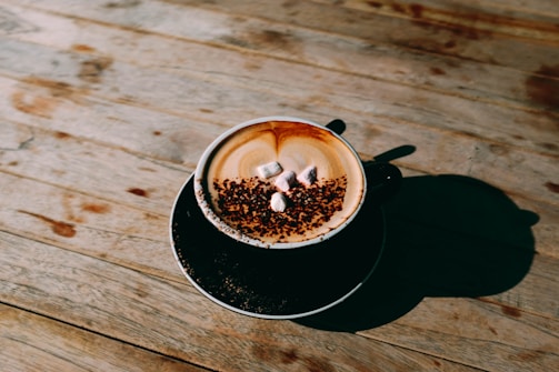A cappuccino with frothed milk and chocolate sprinkles is served in a black cup on a rustic wooden table. Three small marshmallows rest on top of the drink, adding a touch of sweetness. The lighting creates a soft shadow of the cup on the table, enhancing the warm and cozy atmosphere.