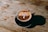 A cappuccino with frothed milk and chocolate sprinkles is served in a black cup on a rustic wooden table. Three small marshmallows rest on top of the drink, adding a touch of sweetness. The lighting creates a soft shadow of the cup on the table, enhancing the warm and cozy atmosphere.