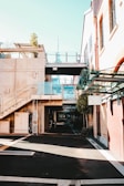 A modern urban area featuring buildings with a blend of concrete and brickwork. Glass railings complement the contemporary design, and a potted plant adds a touch of greenery on the second floor. A distant tower is visible, adding to the cityscape view. The shadows and sunlight create a contrast on the mostly empty pedestrian path.