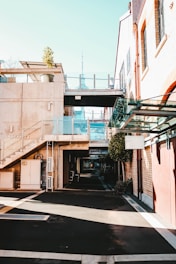 A modern urban area featuring buildings with a blend of concrete and brickwork. Glass railings complement the contemporary design, and a potted plant adds a touch of greenery on the second floor. A distant tower is visible, adding to the cityscape view. The shadows and sunlight create a contrast on the mostly empty pedestrian path.
