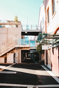 A modern urban area featuring buildings with a blend of concrete and brickwork. Glass railings complement the contemporary design, and a potted plant adds a touch of greenery on the second floor. A distant tower is visible, adding to the cityscape view. The shadows and sunlight create a contrast on the mostly empty pedestrian path.