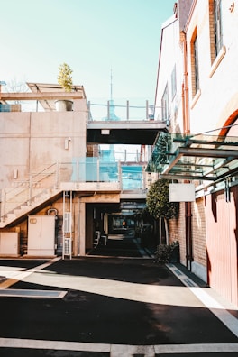 A modern urban area featuring buildings with a blend of concrete and brickwork. Glass railings complement the contemporary design, and a potted plant adds a touch of greenery on the second floor. A distant tower is visible, adding to the cityscape view. The shadows and sunlight create a contrast on the mostly empty pedestrian path.