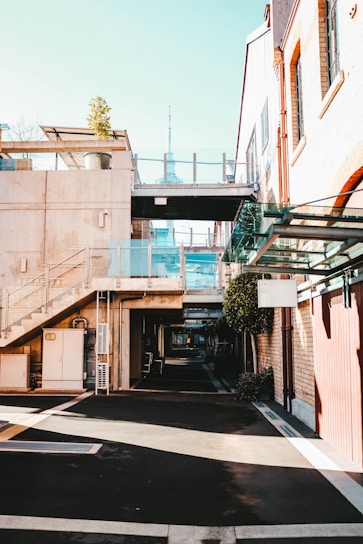 A modern urban area featuring buildings with a blend of concrete and brickwork. Glass railings complement the contemporary design, and a potted plant adds a touch of greenery on the second floor. A distant tower is visible, adding to the cityscape view. The shadows and sunlight create a contrast on the mostly empty pedestrian path.