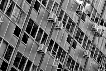 A facade of a building featuring numerous rectangular windows, with several air conditioning units attached to the exterior. The image is in black and white, highlighting the geometric patterns created by the window frames and the orderly placement of the air conditioners.