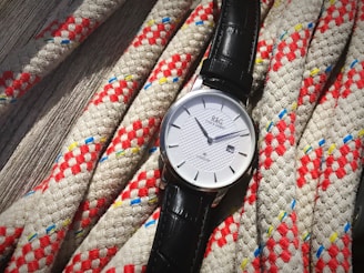 A wristwatch with a white dial and black leather strap rests on intricately woven ropes. The ropes feature a pattern of red and white with occasional blue and yellow threads, creating a textured appearance.